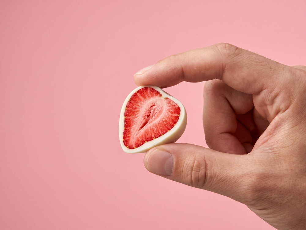 Hand houdt een gehalveerde gevriesdroogde aardbei omhuld met witte chocolade tegen een zachte roze achtergrond