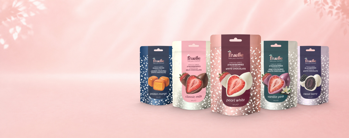 Vijf Fruelle zakjes met gevriesdroogd fruit in chocolade op een zachte roze achtergrond: Golden Mango, Classic Milk, Pearl White, Vanilla Pink en Sweet Berry.