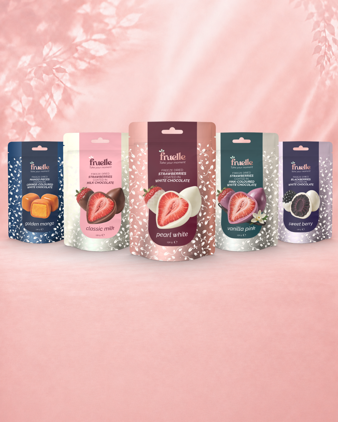 Vijf Fruelle zakjes met gevriesdroogd fruit in chocolade op een zachte roze achtergrond: Golden Mango, Classic Milk, Pearl White, Vanilla Pink en Sweet Berry.